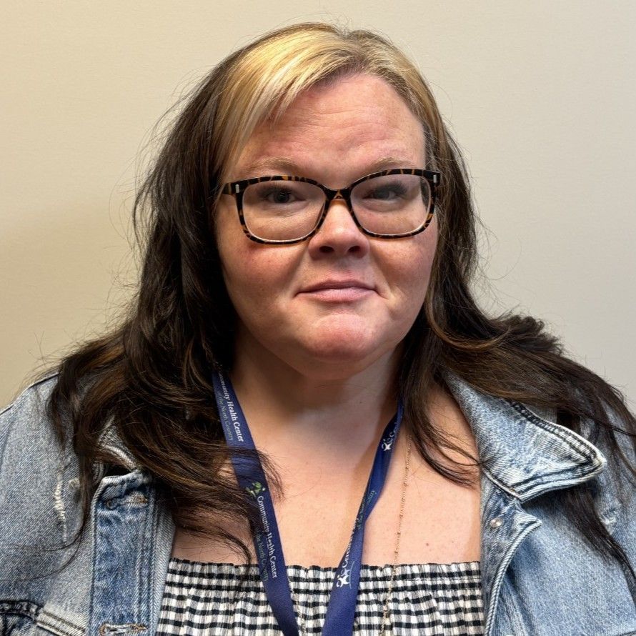 Portrait of an adult person wearing glasses, a denim jacket, and a black-and-white checkered top, standing against a plain light background.