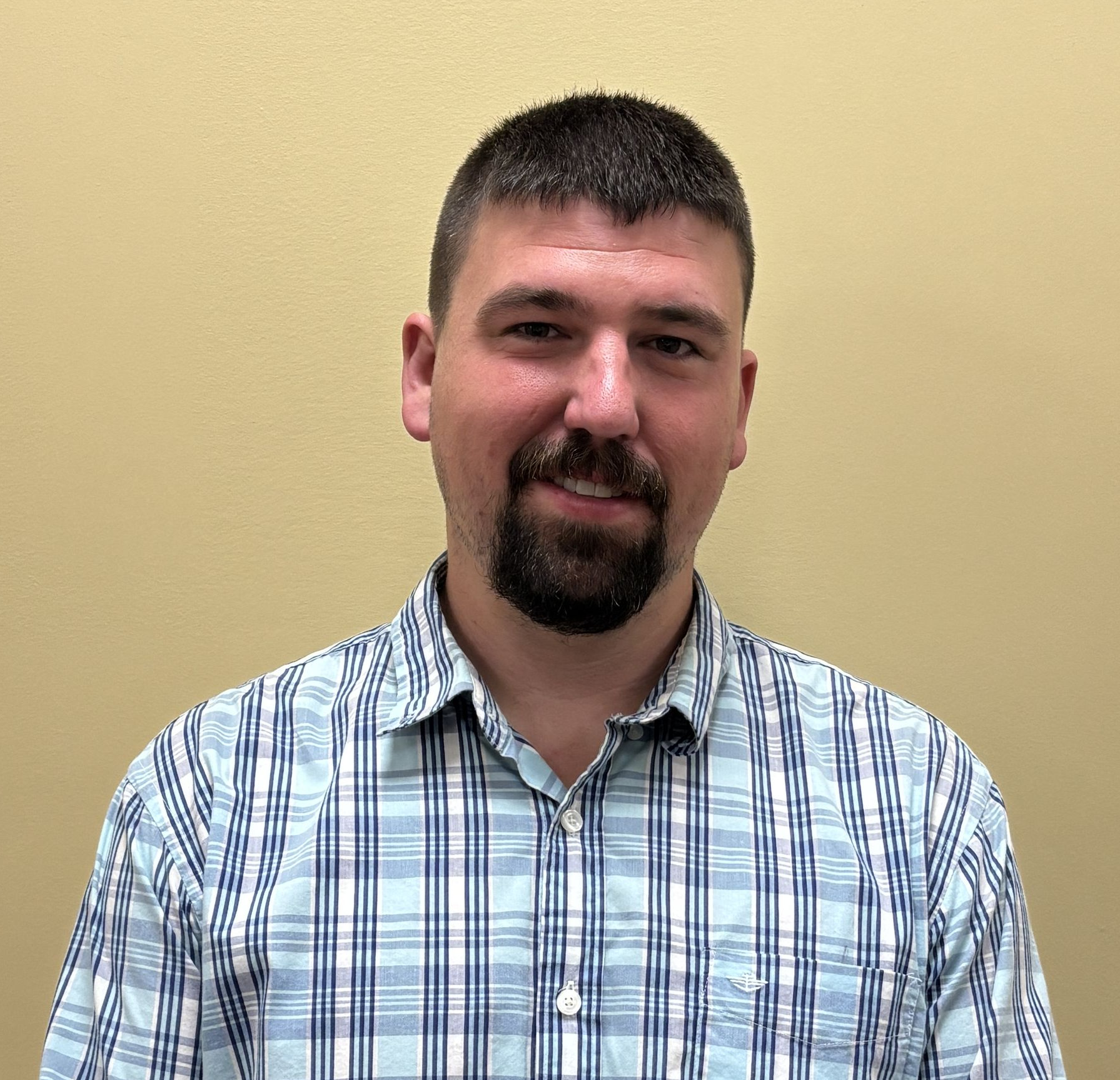 A portrait photograph of a person standing in front of a plain beige wall. The person has short dark hair, a trimmed beard, and is wearing a light blue and white plaid button‑up shirt. The lighting is even, and the image appears to be taken indoors.