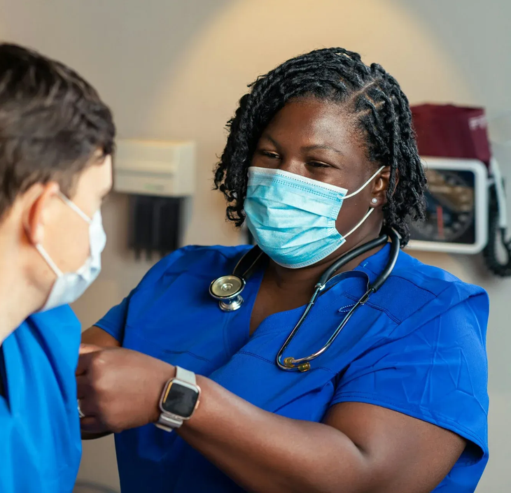 Nurse in blue scrubs and mask adjusts a patient’s arm in a clinic exam room.