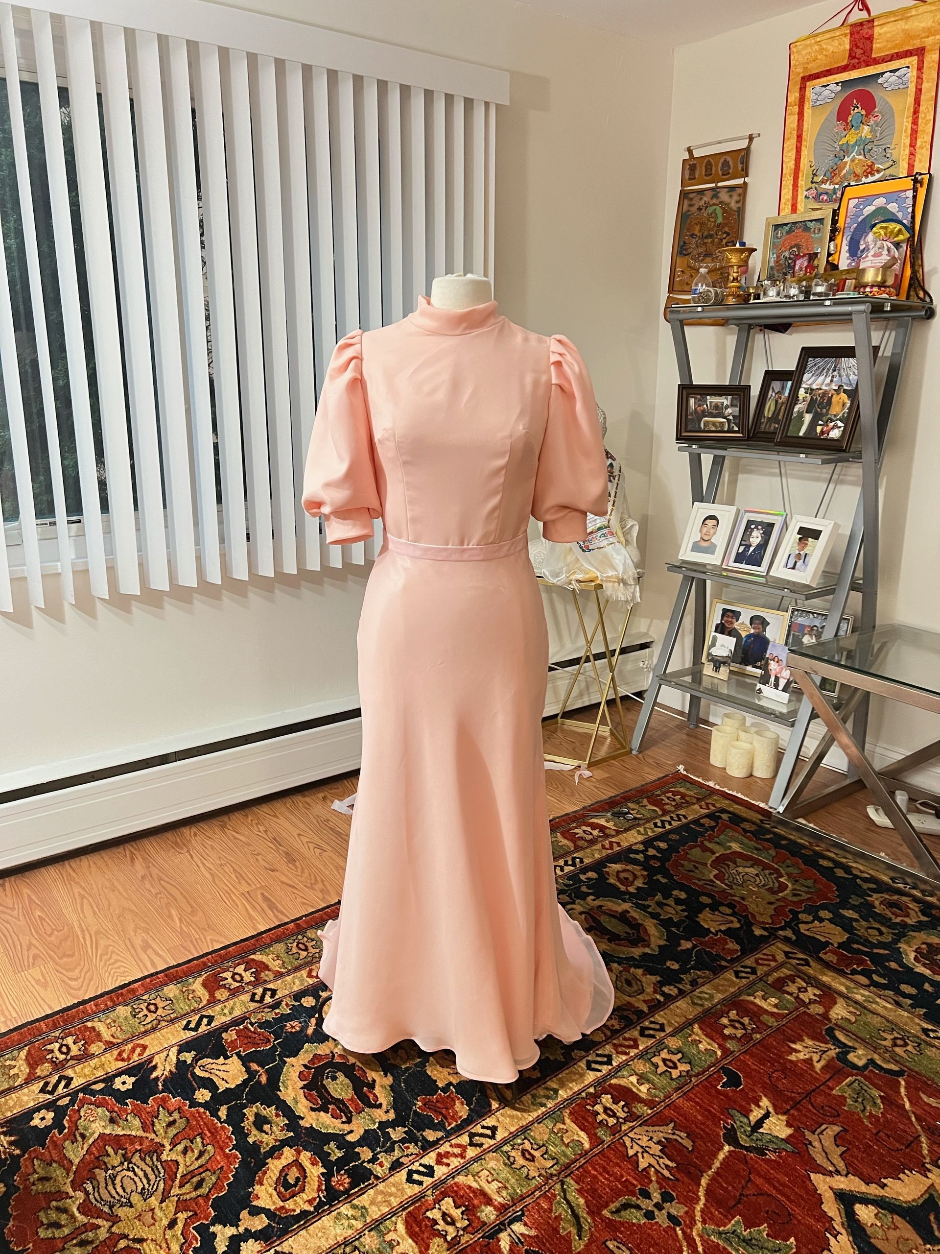 Pale pink dress on a mannequin; puff sleeves and floor-length skirt. Set indoors on a rug, near a window.