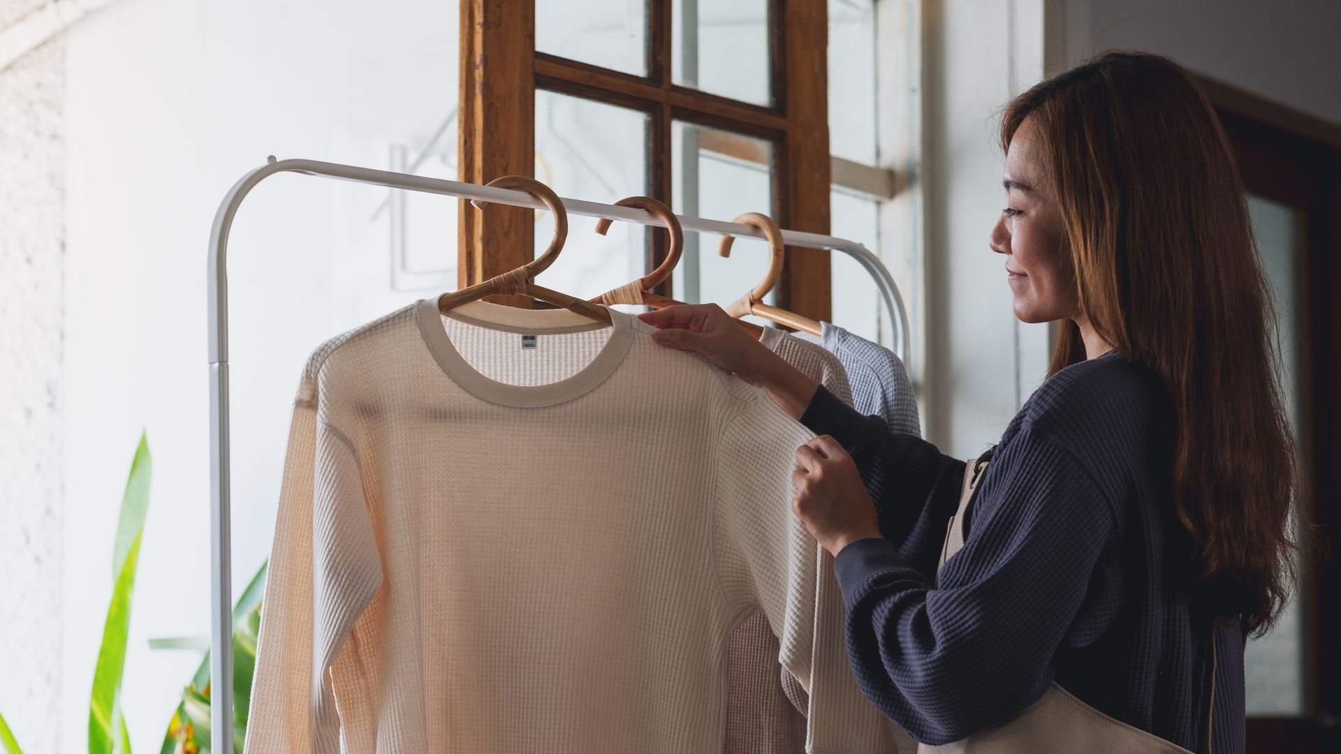 Woman selects a sweater from a clothing rack near a window.