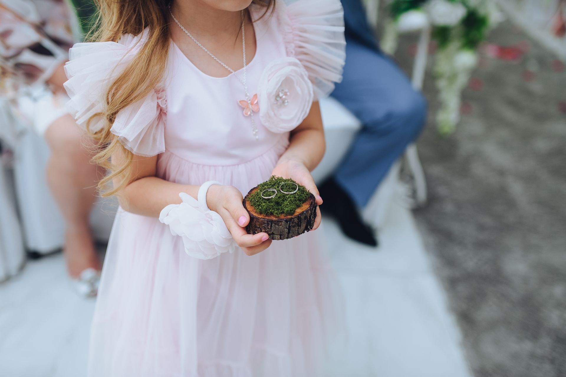 Flower girl in pink dress holding a ring box covered in moss at an outdoor wedding.
