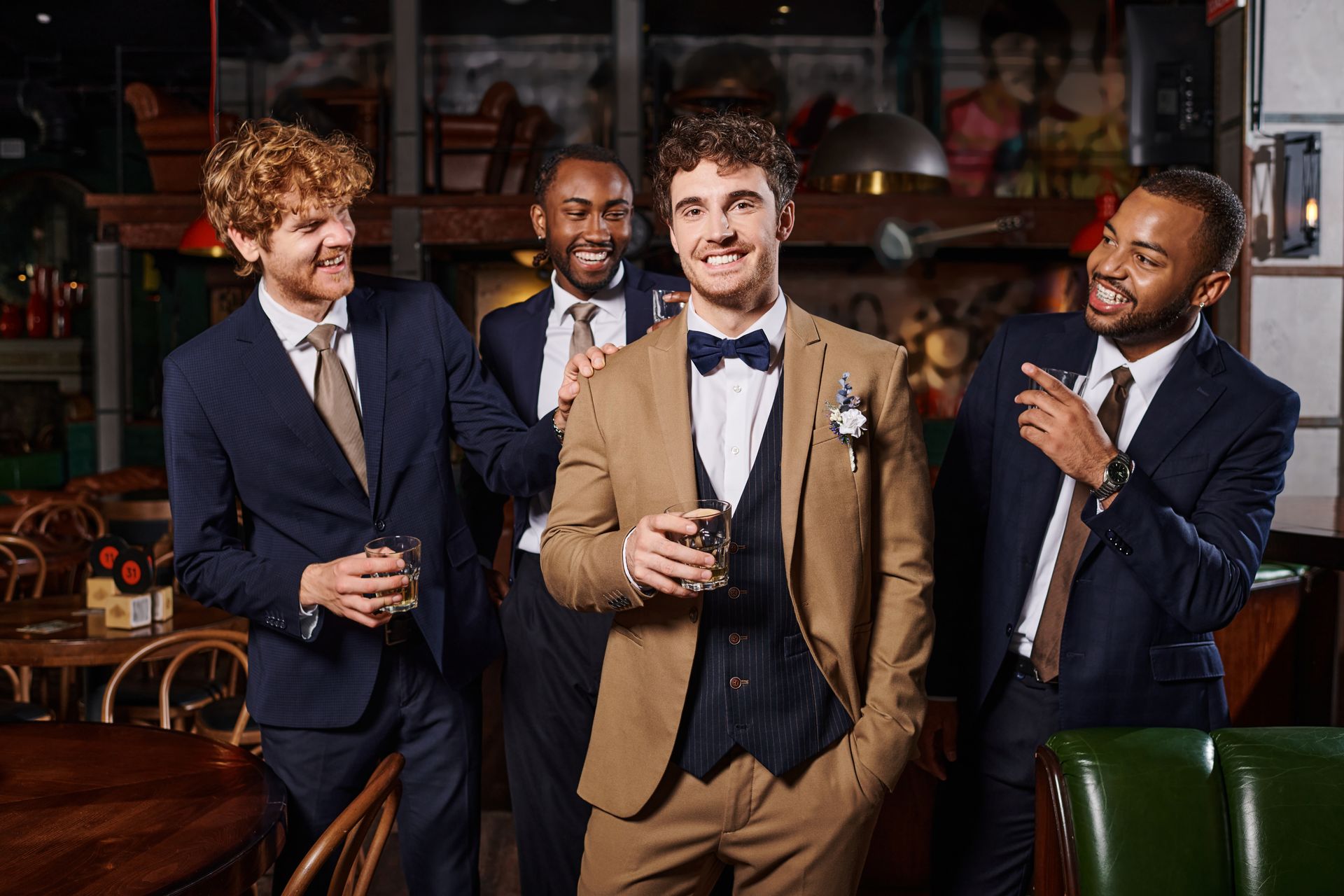 Four men in suits at a bar, toasting with drinks. The man in the tan suit smiles.