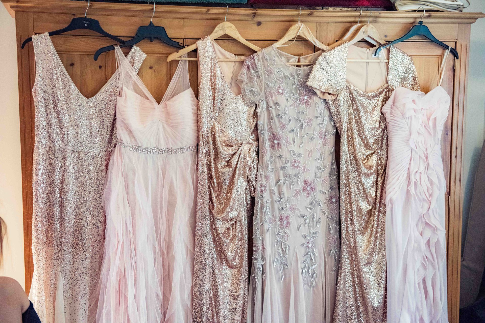 Six sequined and flowing bridesmaid dresses in shades of pink and gold hanging in a closet.