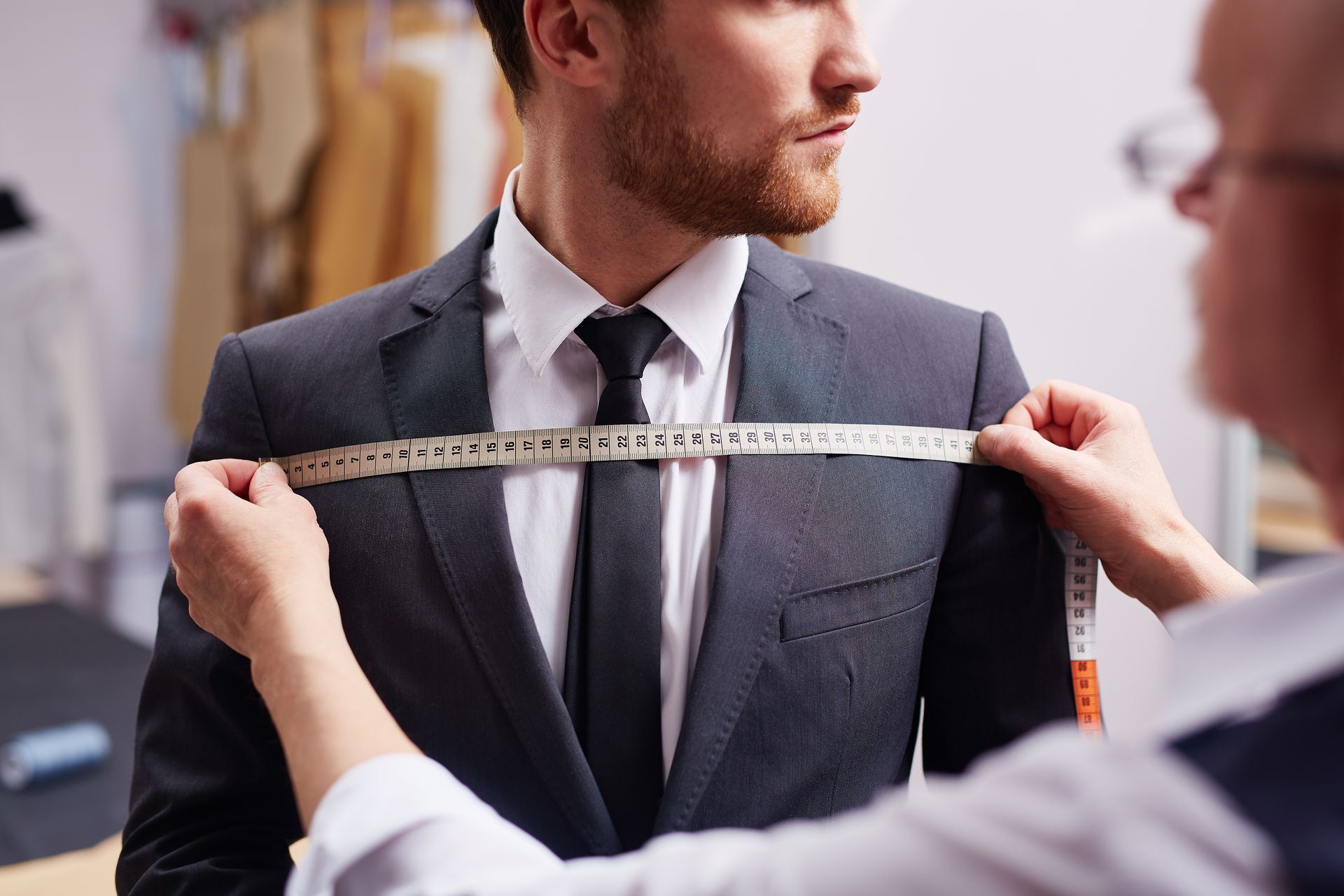 Tailor measuring a man's shoulders for a suit fitting. White measuring tape and collar visible.