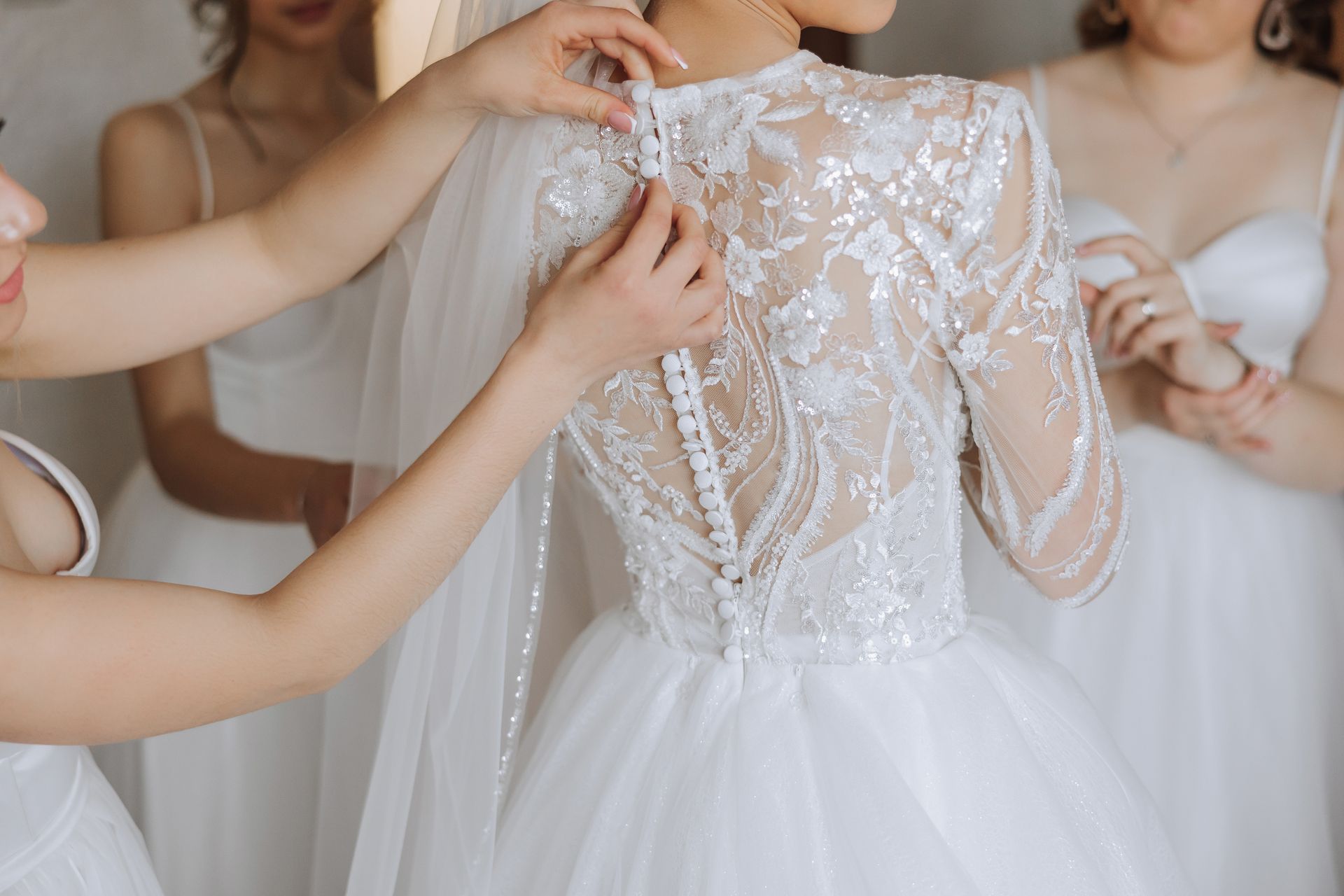 Bridesmaids helping the bride fasten buttons on the back of her white lace wedding dress.
