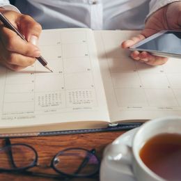 A person is writing in a planner while holding a cell phone