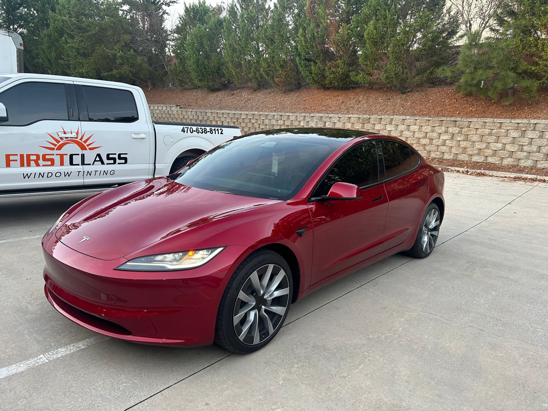 A red tesla model 3 is parked in a parking lot next to a white truck.
