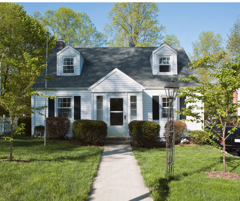 White Cape Cod home with black shutters, a gray roof, and a concrete walkway, set on a green lawn.