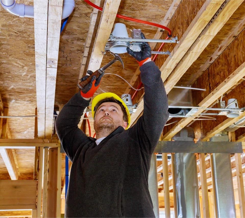 Construction worker hammering a light fixture into a wooden ceiling, wearing a yellow hard hat and gloves.