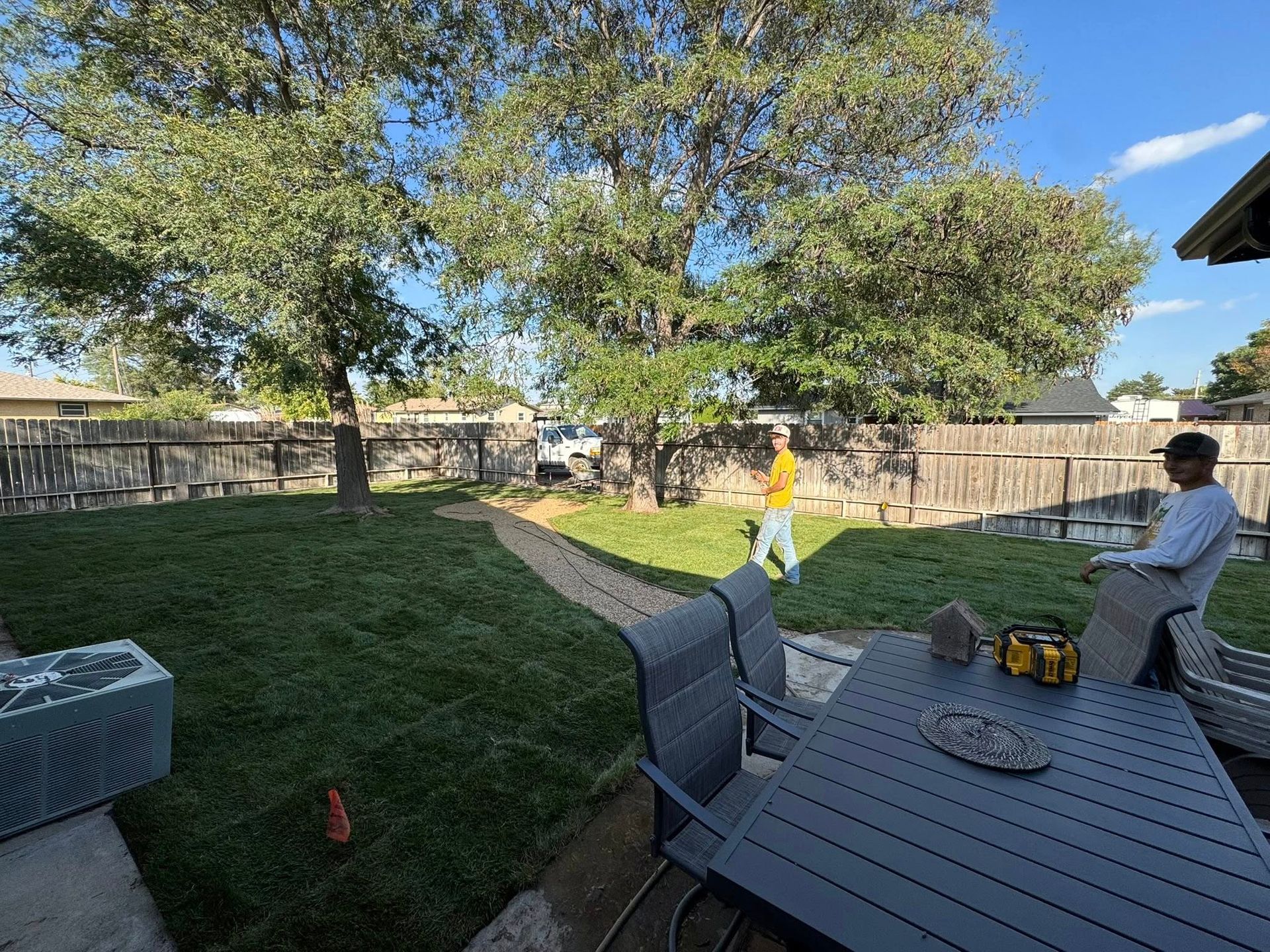 Backyard with patio table, trees, fence, and two people relaxing in the yard