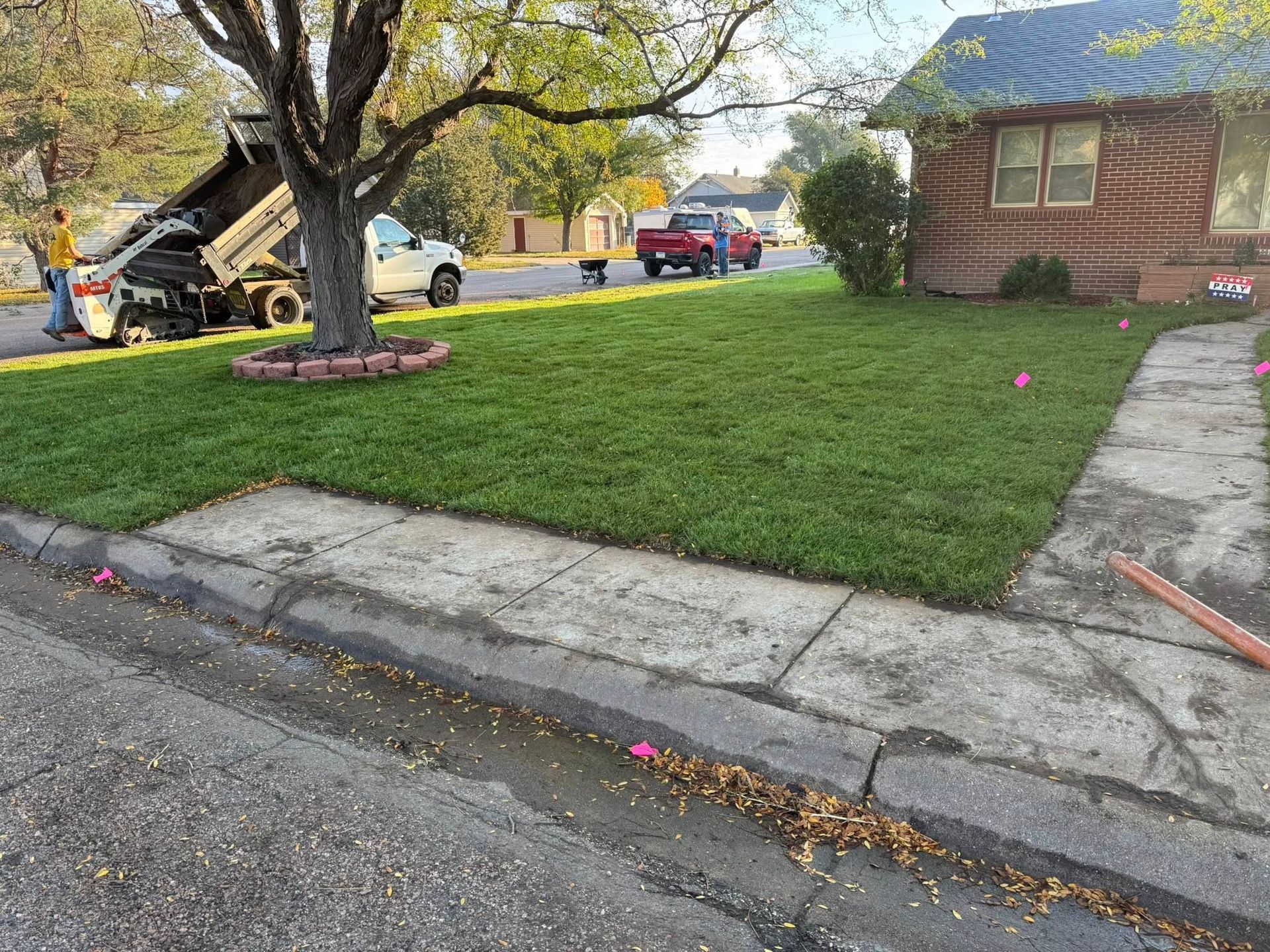 Suburban yard with a lawn, tree, sidewalk, parked cars, and a utility truck near a brick house.