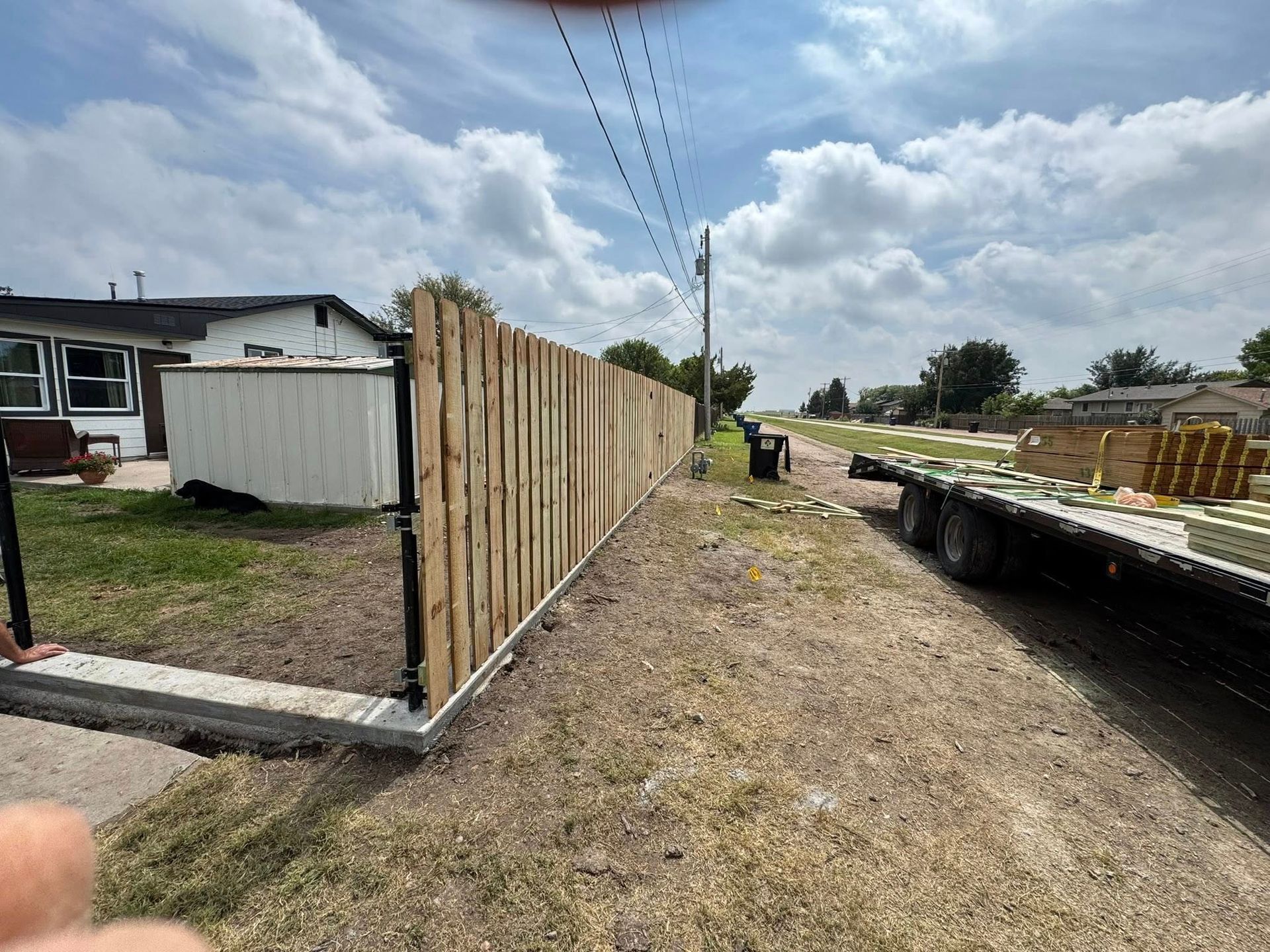 Residential construction site with a wooden fence along a dirt path beside a flatbed truck under a cloudy sky