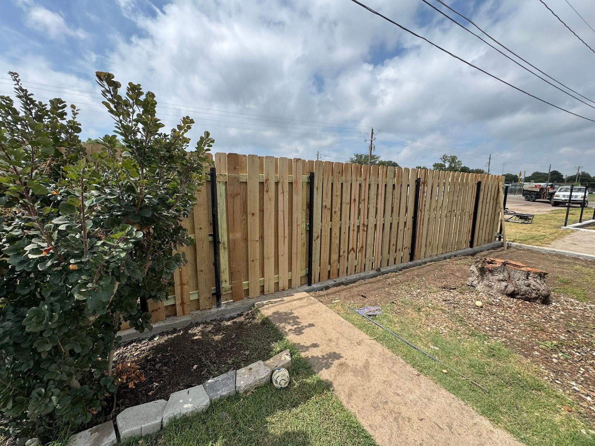 Wooden fence along a dirt path under a cloudy sky, with shrubs and a driveway nearby