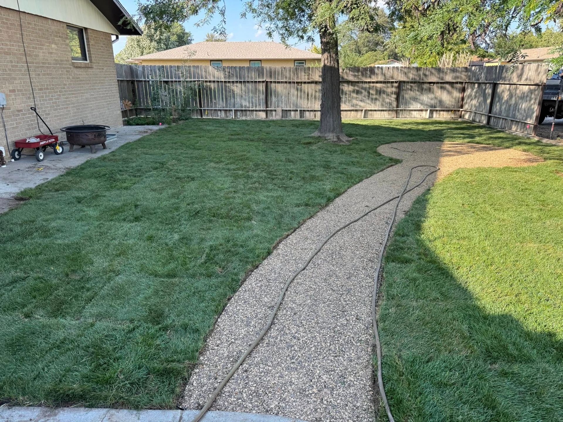 Backyard with a curved gravel path, green lawn, wooden fence, and trees in sunlight