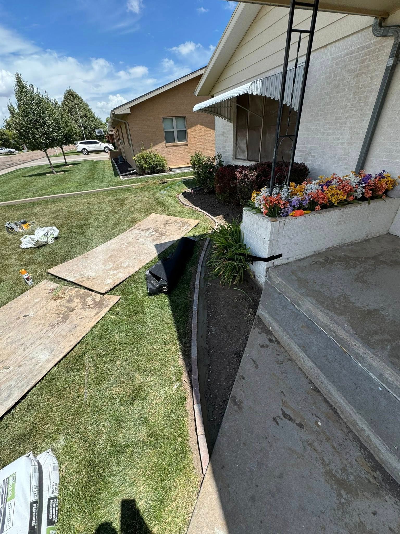 Damaged sidewalk with a deep trench beside a house, flower planter, and boards laid across grass