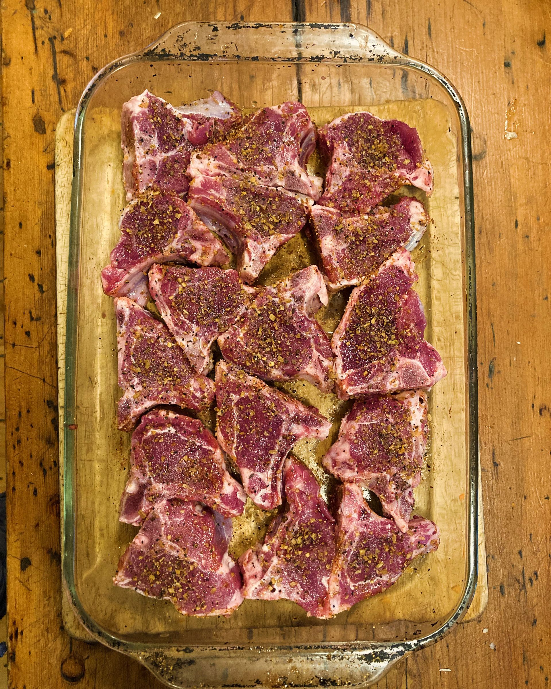 Raw, seasoned lamb chops arranged in a glass baking dish, on a wooden surface.