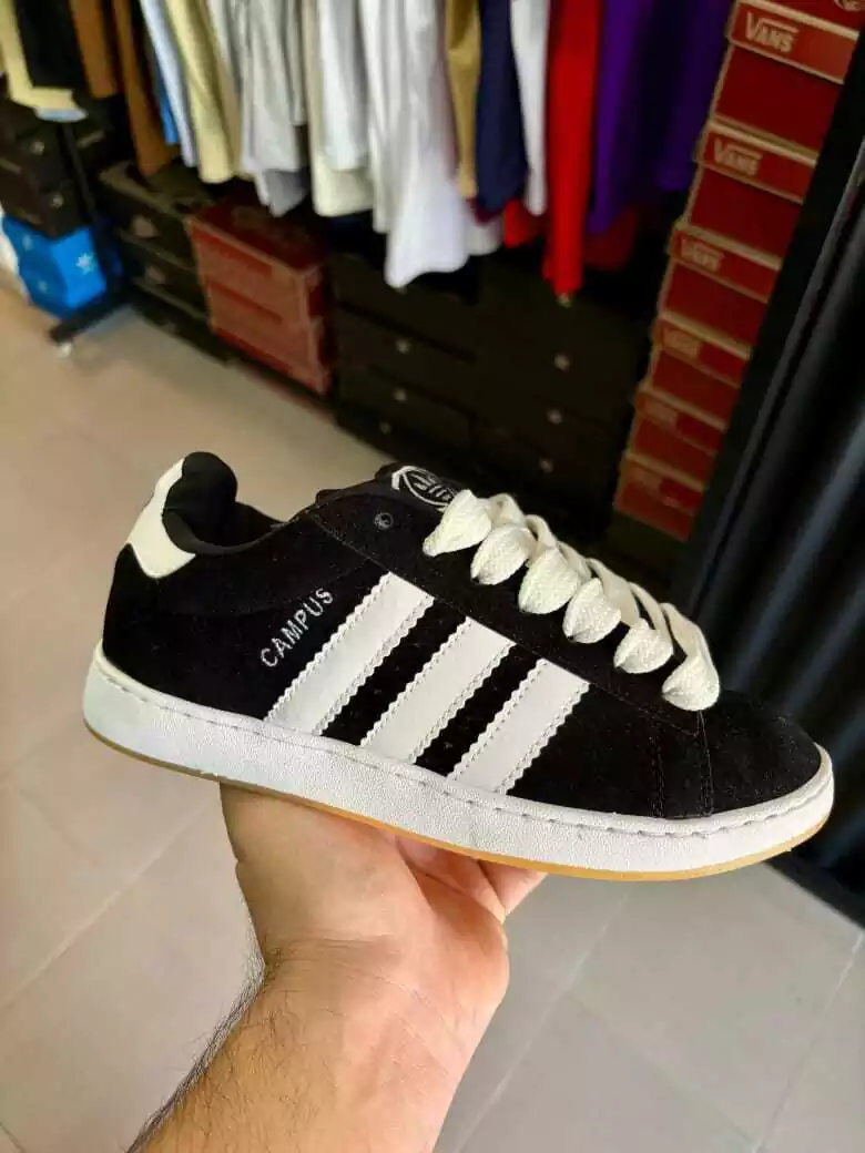 Mano sosteniendo una zapatilla Adidas Campus negra con rayas, cordones y suela blancos. 