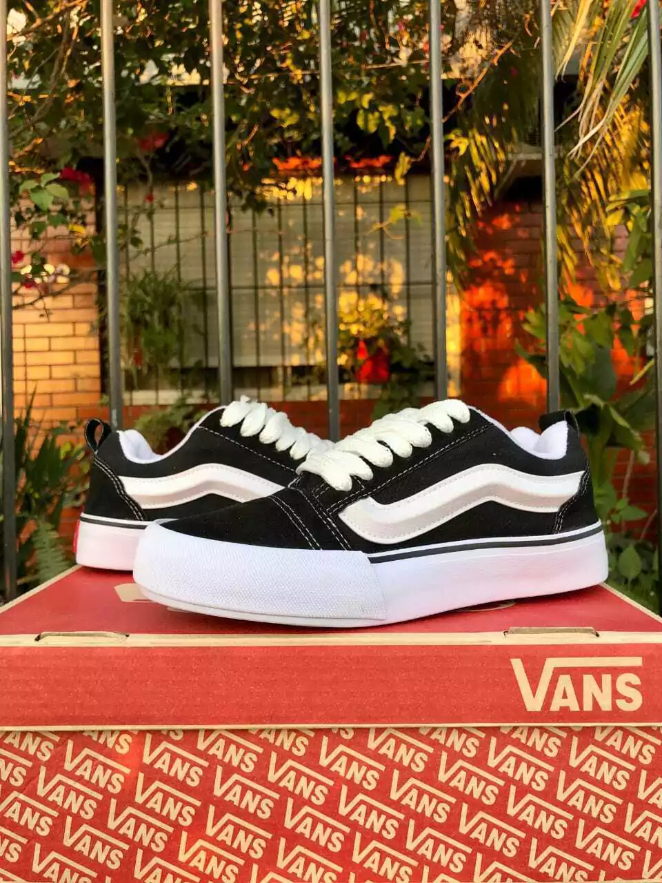 Zapatillas Vans en blanco y negro con suela gruesa blanca, apoyadas sobre una caja de Vans frente a una valla y vegetación.