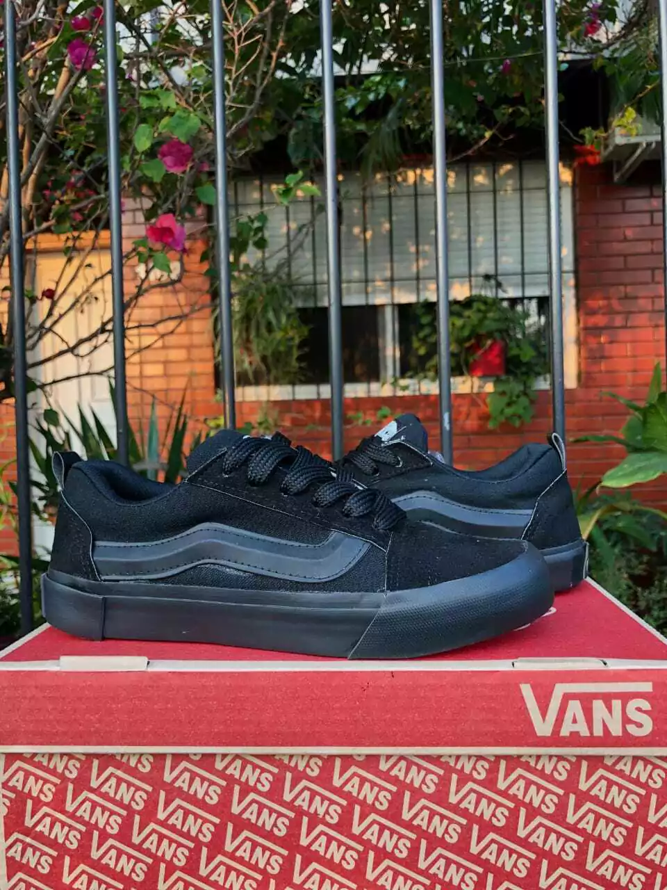 Zapatillas Vans negras sobre una caja de Vans con un edificio de ladrillos y una valla de metal al fondo.