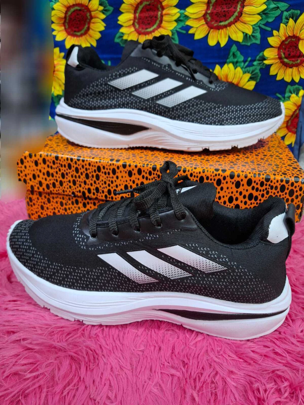 Dos zapatillas Adidas en color blanco y negro con tres rayas plateadas, sobre una superficie rosa y una caja estampada.