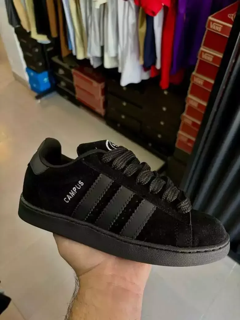 Zapatilla Adidas Campus negra en la mano, colocada en una tienda de ropa con prendas de vestir al fondo.