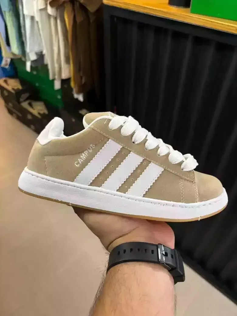 Zapatilla Adidas Campus en color beige con rayas y cordones blancos, sujeta en una mano.