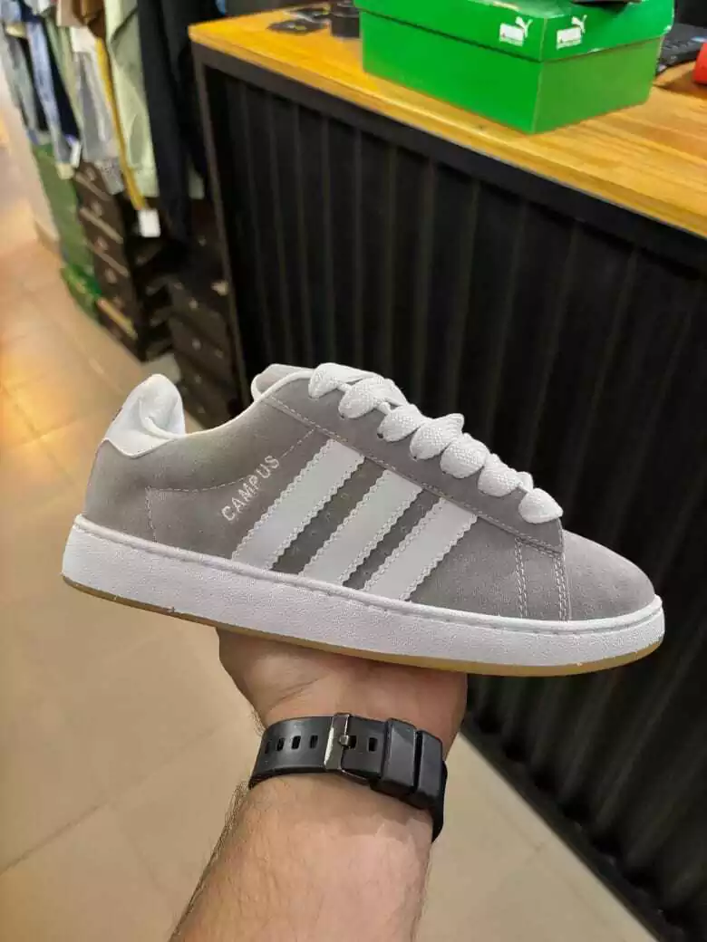 Zapatilla Adidas Campus de color gris con rayas blancas, sostenida en la mano, en interior.