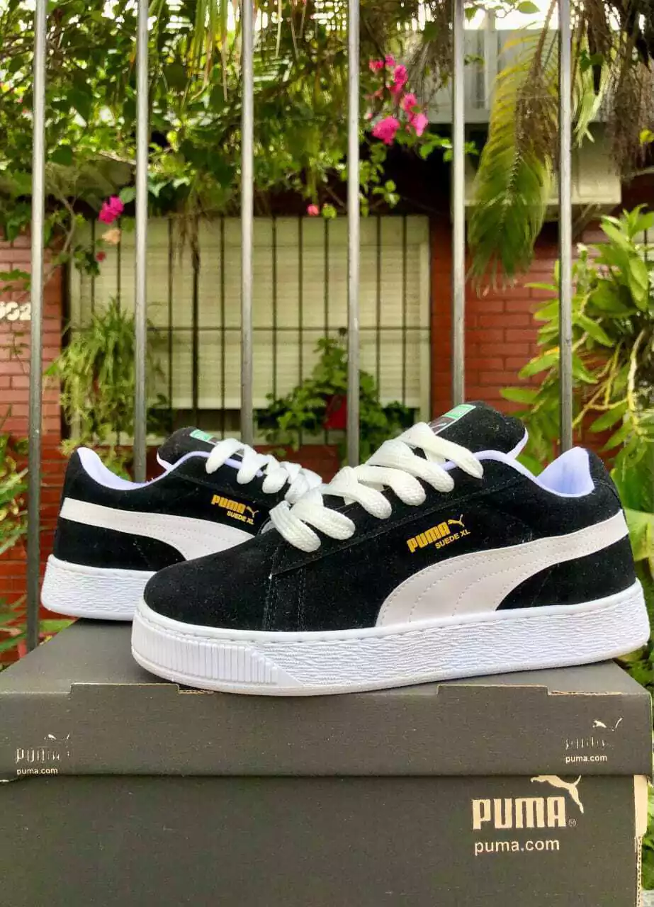 Zapatillas Puma de ante negras con suela, rayas y cordones blancos, sobre una caja Puma gris.