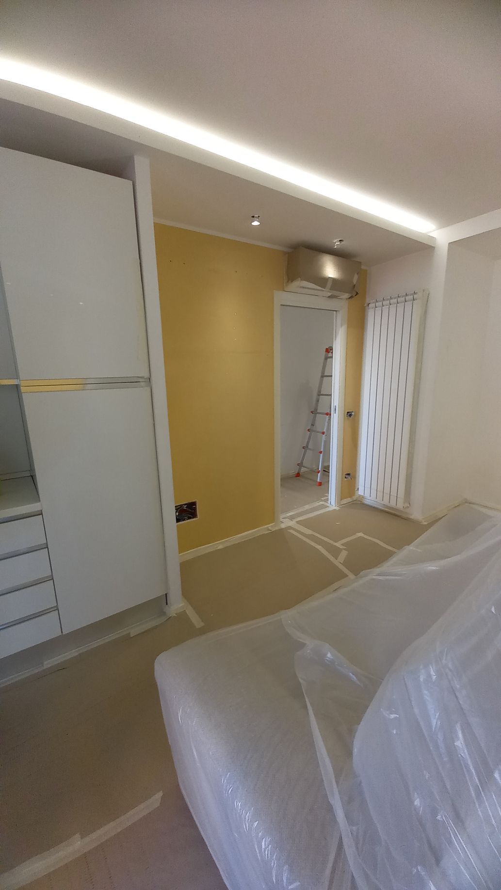 Camera da letto in fase di ristrutturazione con mobili, scala e armadio ricoperti di plastica, addossati a una parete gialla.
