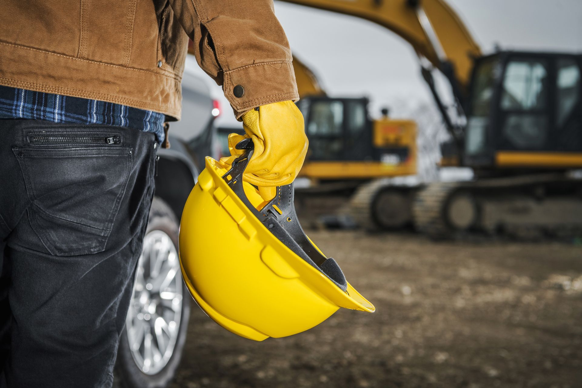 Construction worker in brown jacket holding yellow hard hat, excavator in background.