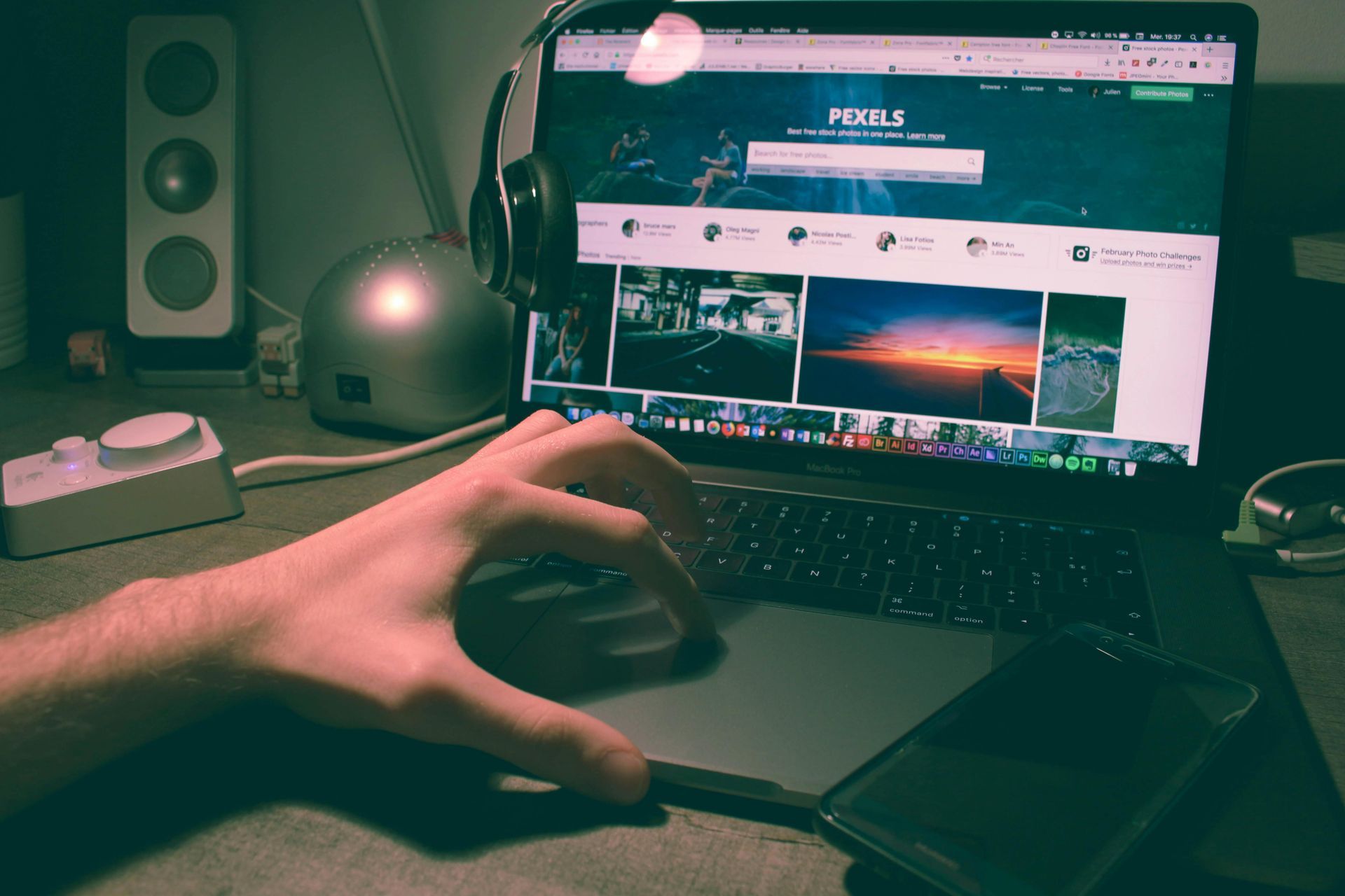 Hand resting on a laptop trackpad in a dimly lit workspace with a computer monitor, headphones, and a speaker.