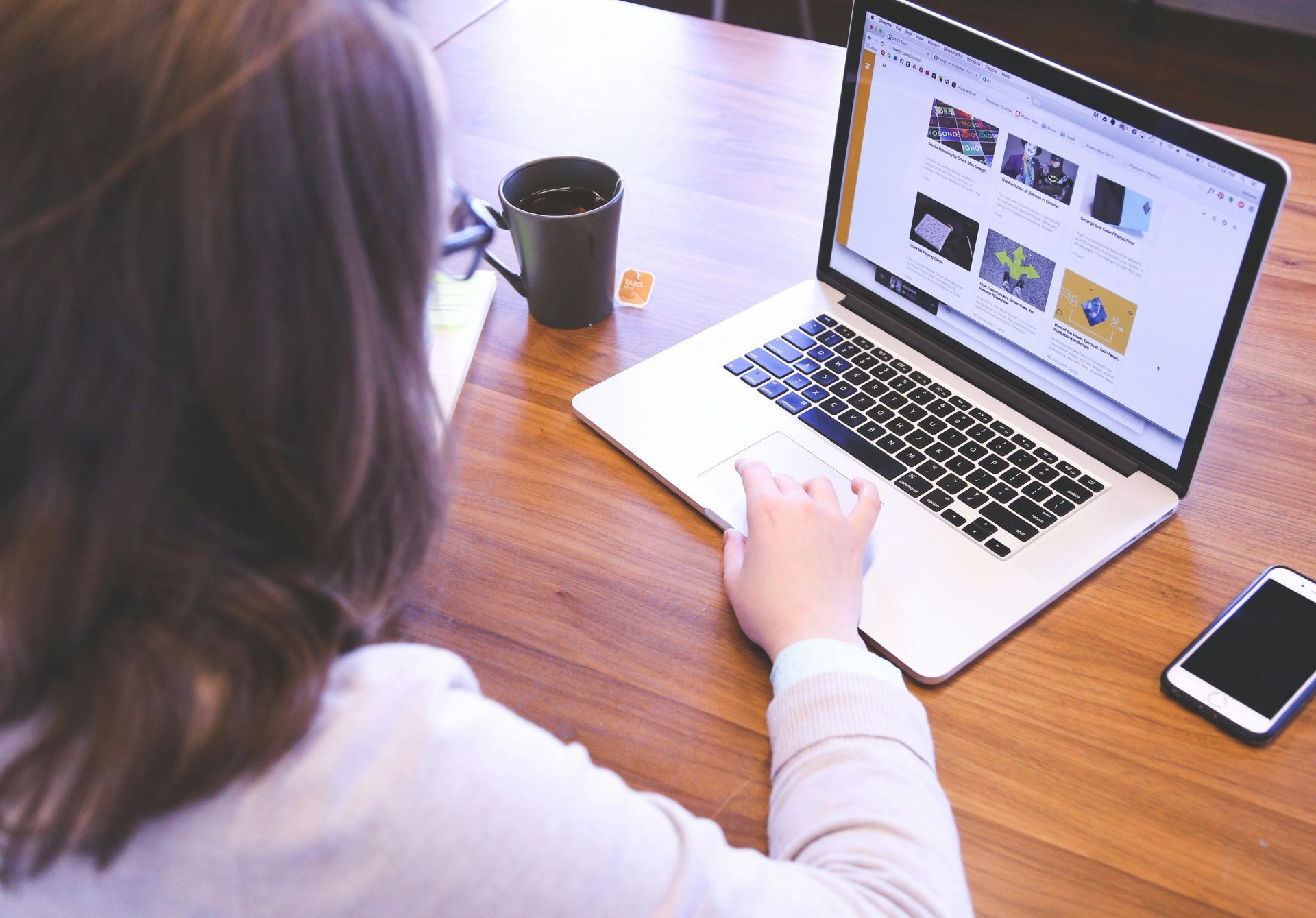 Person at a wooden desk working on a laptop with a mug and smartphone nearby.