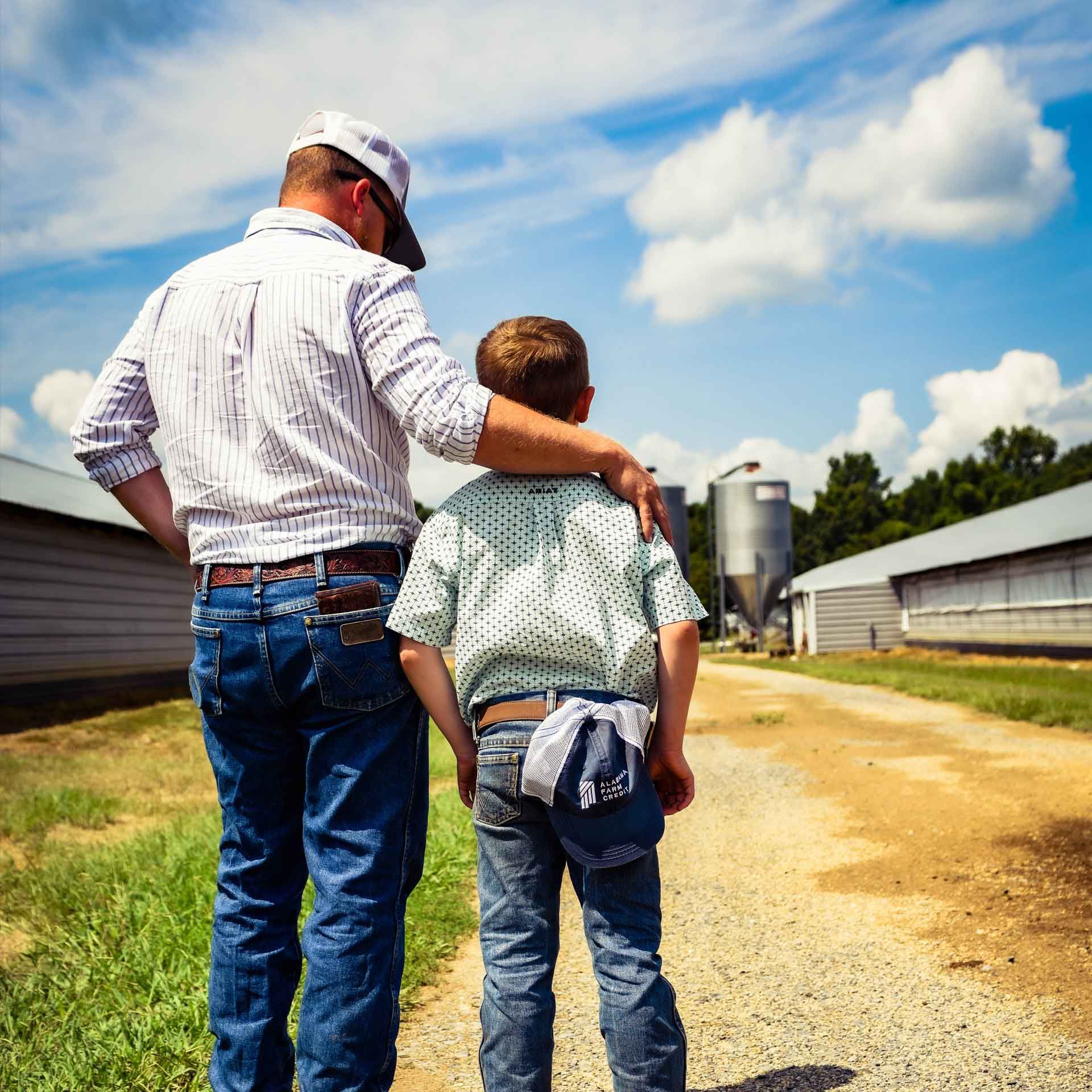 Man with arm around a boy's shoulder walking down a farm path past outbuildings on a sunny day.