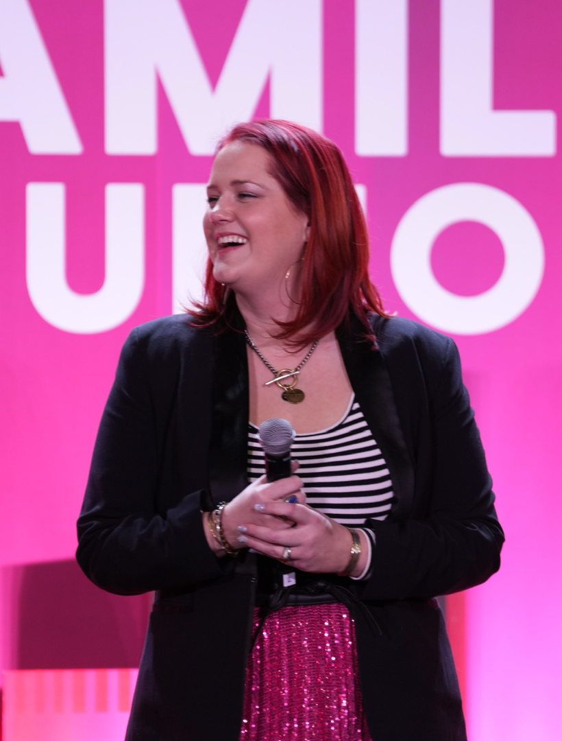 Woman with red hair, holding a microphone, smiling, on a pink stage. Wearing a black blazer, striped shirt, and pink skirt.