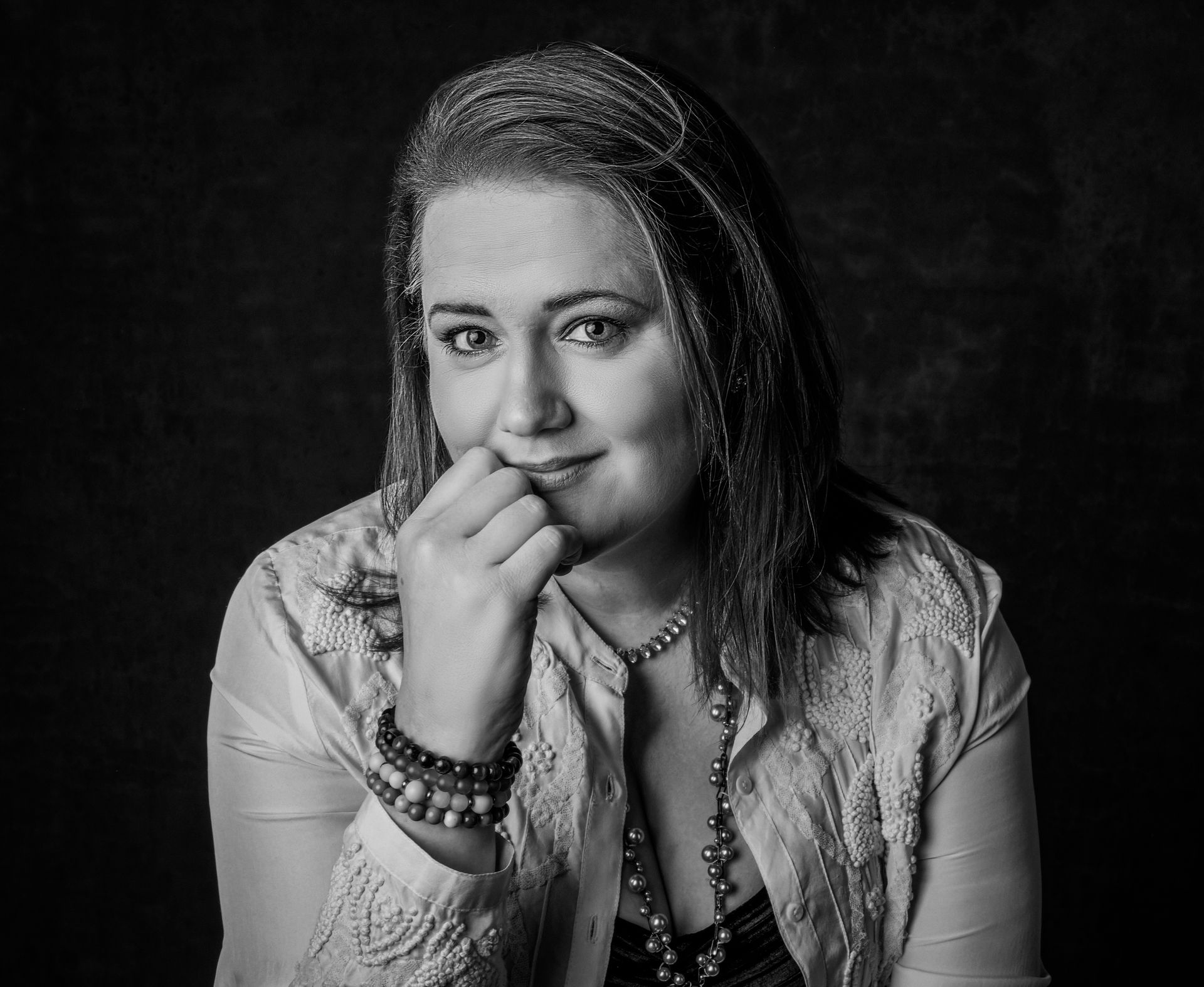 Woman with hand on chin, wearing beaded jewelry, smiling, against a dark backdrop.