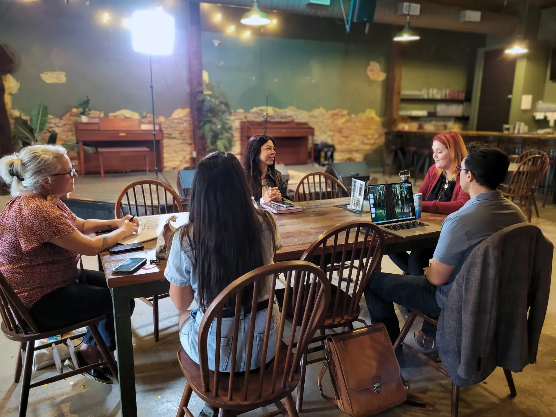 Woman with hand on chin, wearing beaded jewelry, smiling, against a dark backdrop.