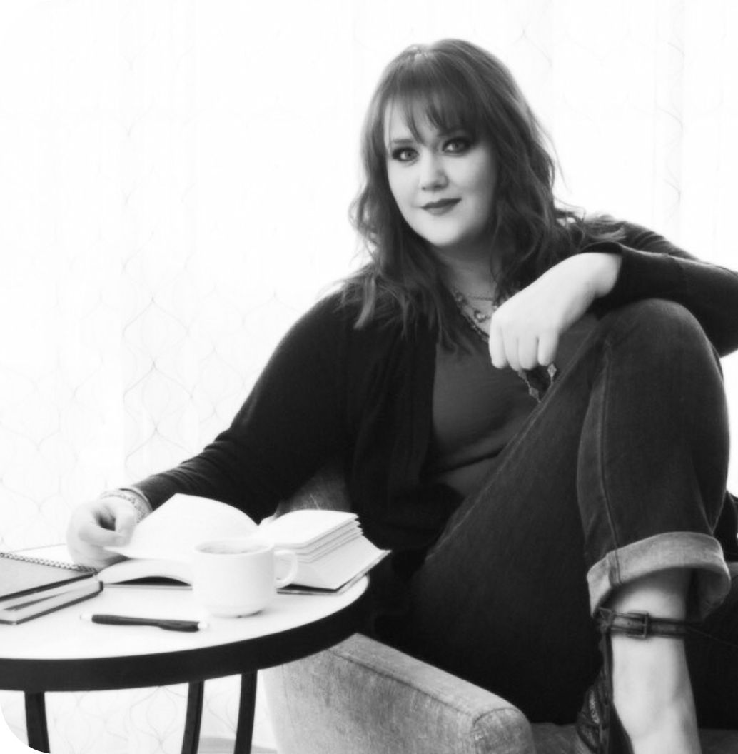 Woman sitting, leaning on a small table with books and a coffee cup. Wearing jeans and a cardigan.