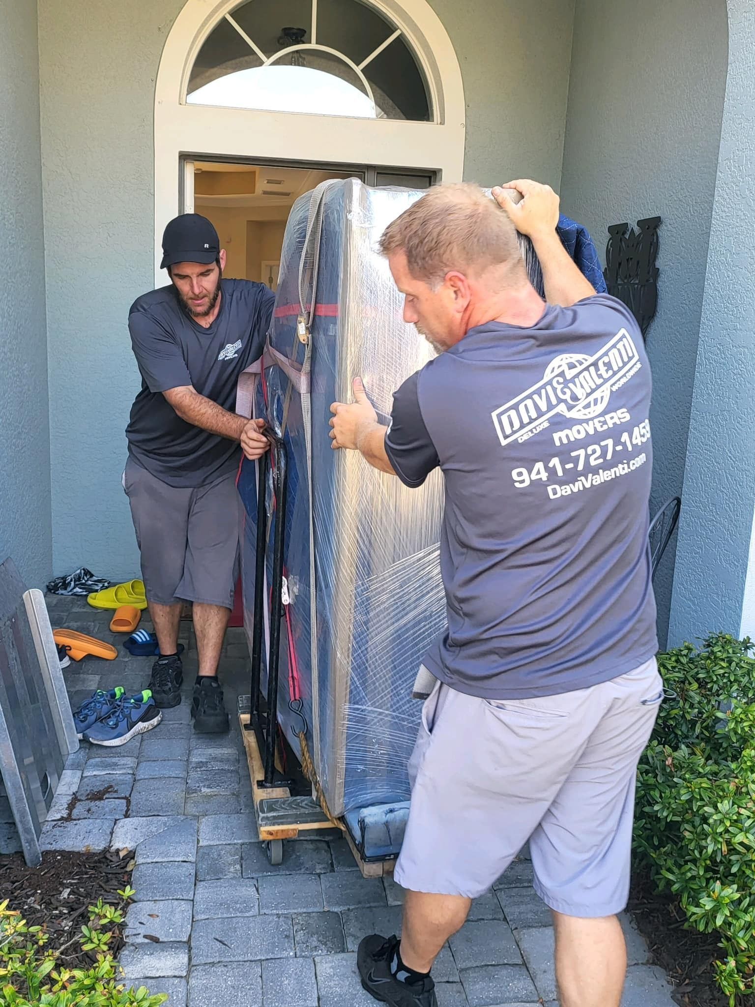 Two movers in navy shirts carrying a wrapped sofa through a front doorway.