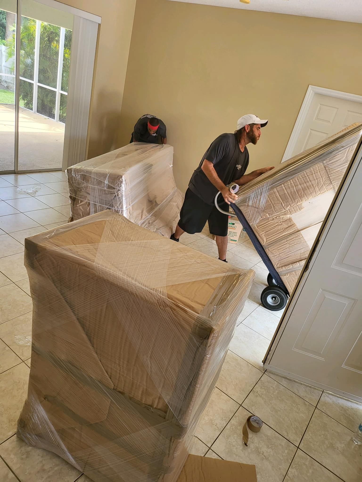 Man using a dolly to move a door, with wrapped furniture in a room.