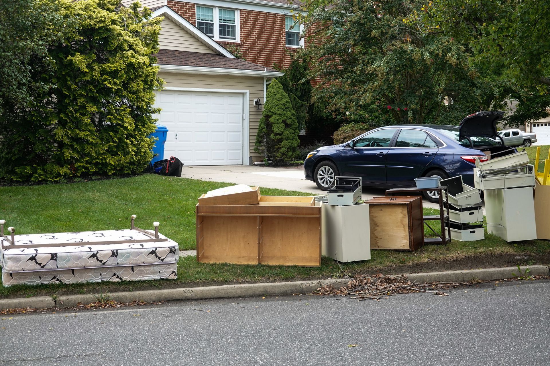 Assorted household furniture and items placed on the lawn near the curb of a suburban house for trash collection.
