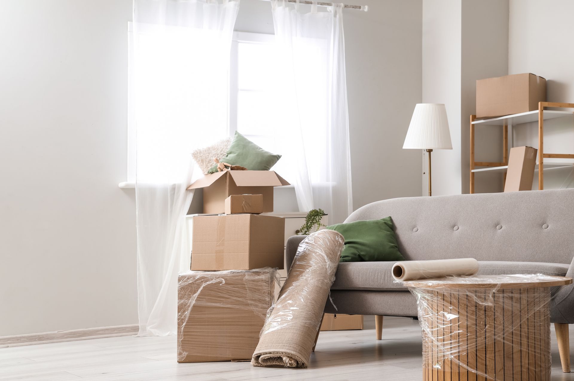 Boxes and wrapped items in a living room, ready for a move, next to a sofa and window.