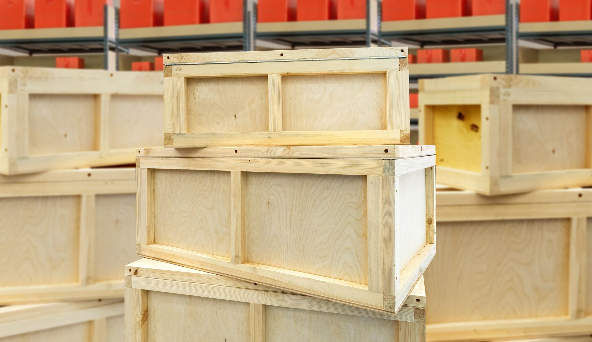 A stack of light-colored wooden crates in a warehouse with rows of red containers on shelves in the background.