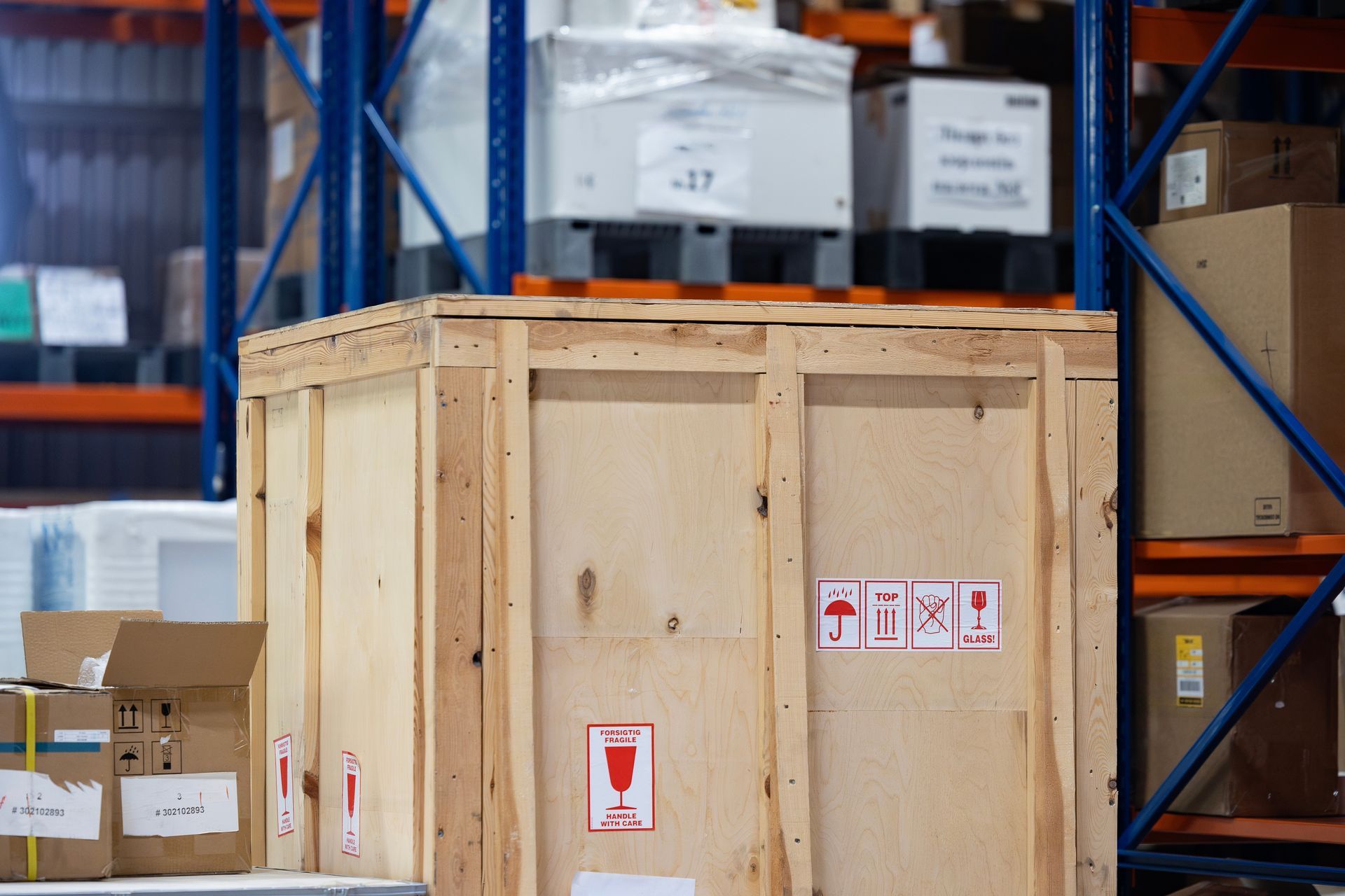 A large wooden shipping crate marked with fragile icons sits on a warehouse shelf alongside various cardboard boxes.