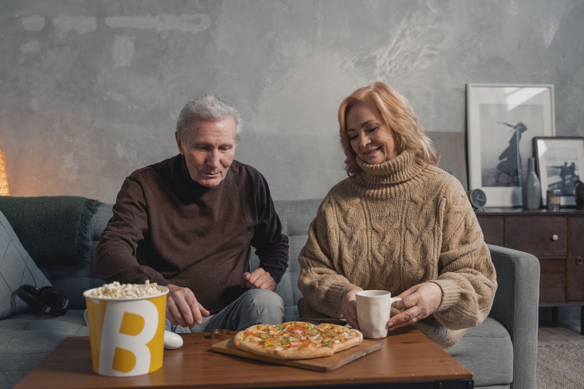 A couple are sitting on their couch looking at a pizza and box of popcorn on their coffee table. They are both wearing earth-toned sweaters.