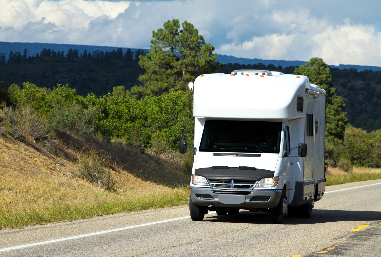 White RV driving on a paved road, mountains and trees in the background under a blue and cloudy sky.