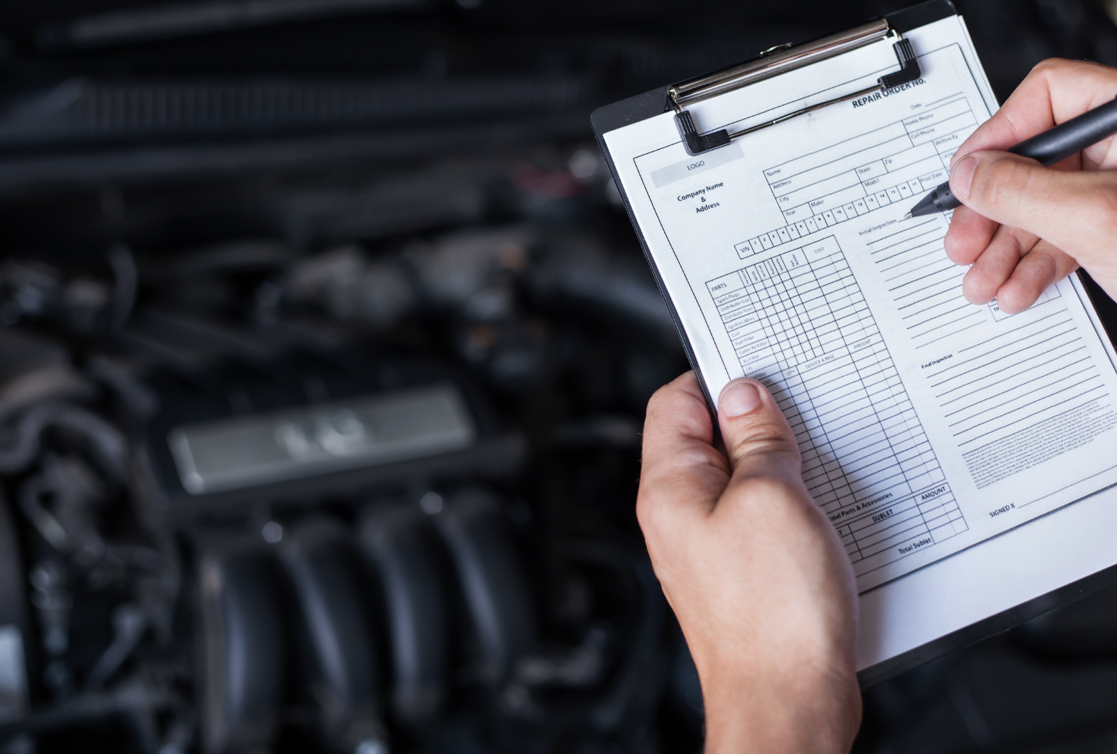 Mechanic's hands holding clipboard, writing notes near car engine.