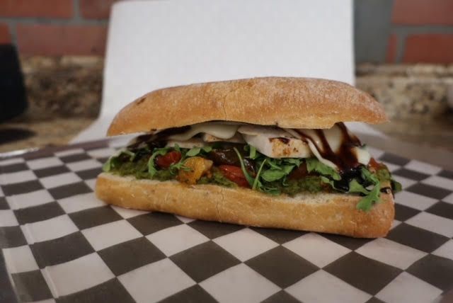 A sub sandwich is sitting on a checkered paper next to a cup of dipping sauce.