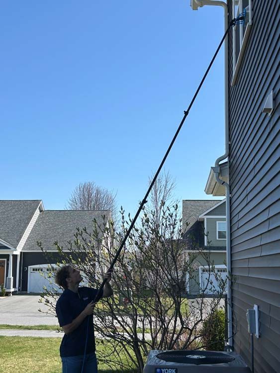 Person cleaning gutters with an extended pole on a residential house exterior; blue sky background.