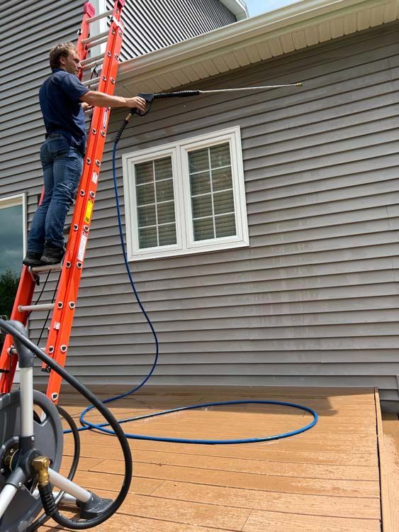Man on a ladder pressure washing a gray house siding near a window and deck